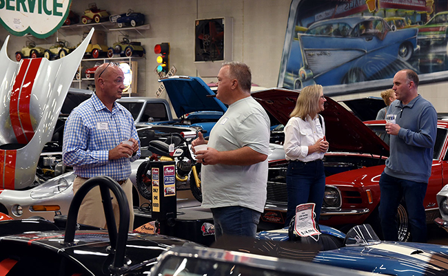 Four advisors stand and talk inside a classic car showroom, surrounded by vintage cars with open hoods and bright paint jobs. Two men chat near the left side, while a man and woman talk near a red car on the right. The background features toy cars on shelves, a large wall mural of classic vehicles, and retro signage.