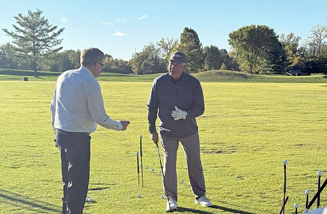 Two men stand on a sunny golf course, talking near a line of golf balls on the grass. One man gestures with his hand while the other, wearing a cap and golf glove, holds a club. Trees and blue sky frame the background