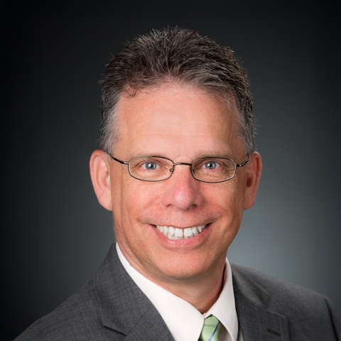 Headshot of Dave Kaplan, wearing a white shirt, green tie, and gray blazer, with a plain gray background