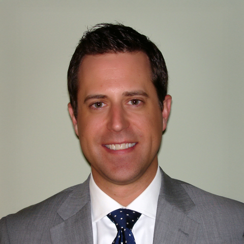 Headshot of Mark Beckrich, wearing a white shirt, blue tie, and gray blazer, with a plain gray background