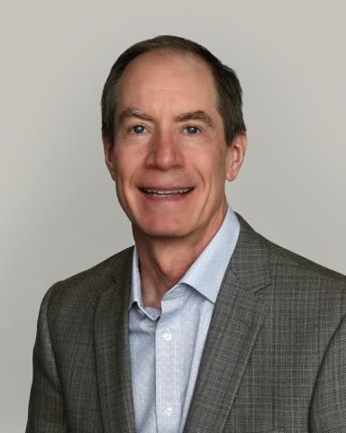Headshot of Tom Gooden, wearing a light shirt and gray blazer, with a plain gray background