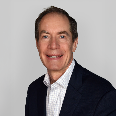 Headshot of Tom Gooden, wearing a white shirt and blue blazer, with a plain gray background