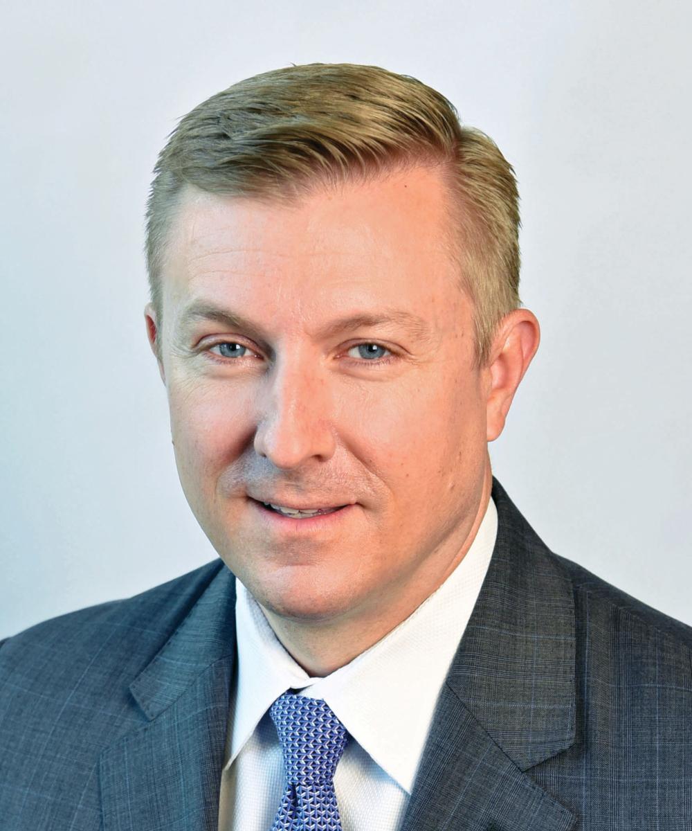Headshot of Bill Friebel, wearing a white shirt, blue patterned tie, and gray blazer, with a plain gray background