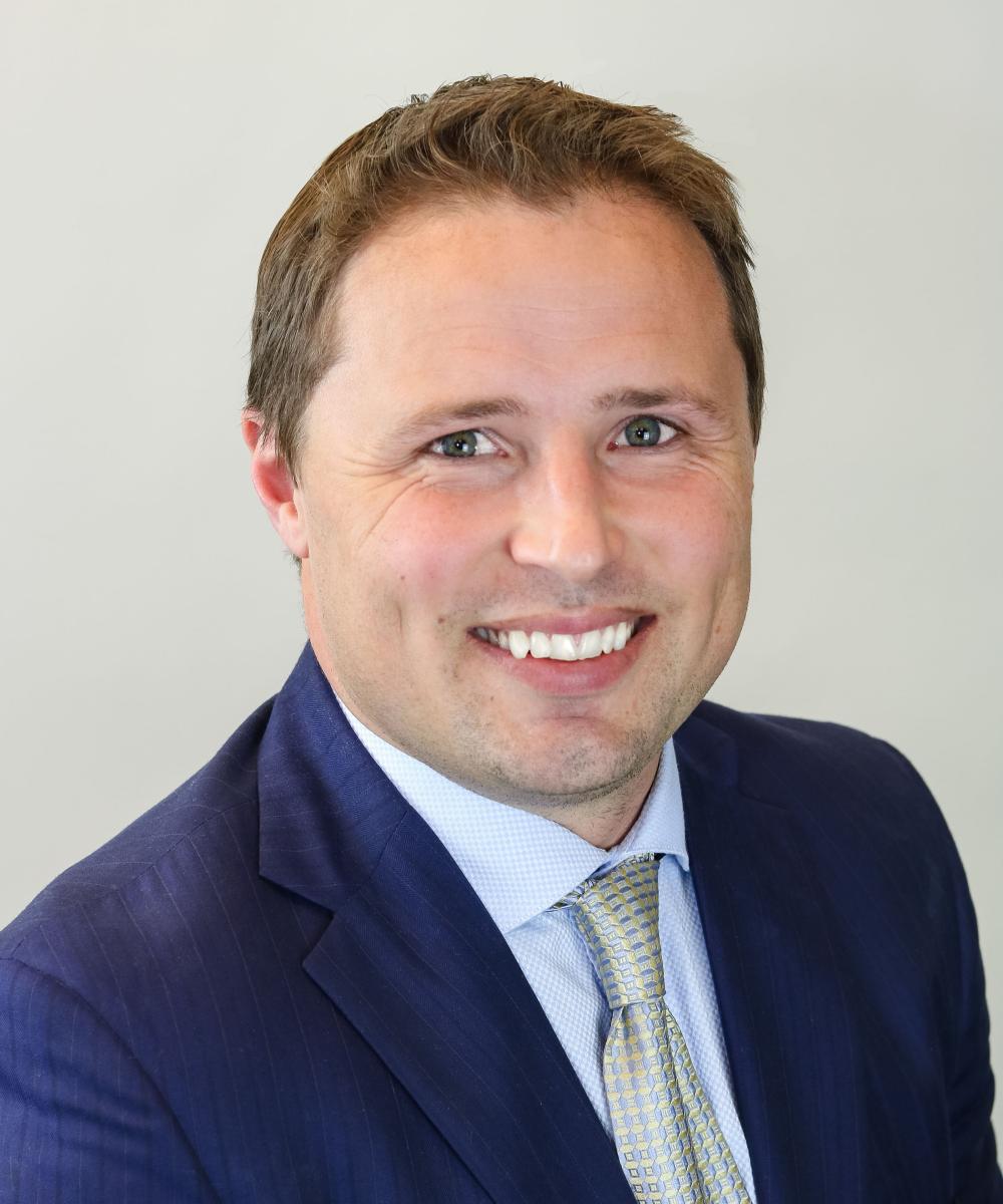 Headshot of Casey Loper wearing a light blue shirt, gray tie, and blue blazer, with a plain gray background