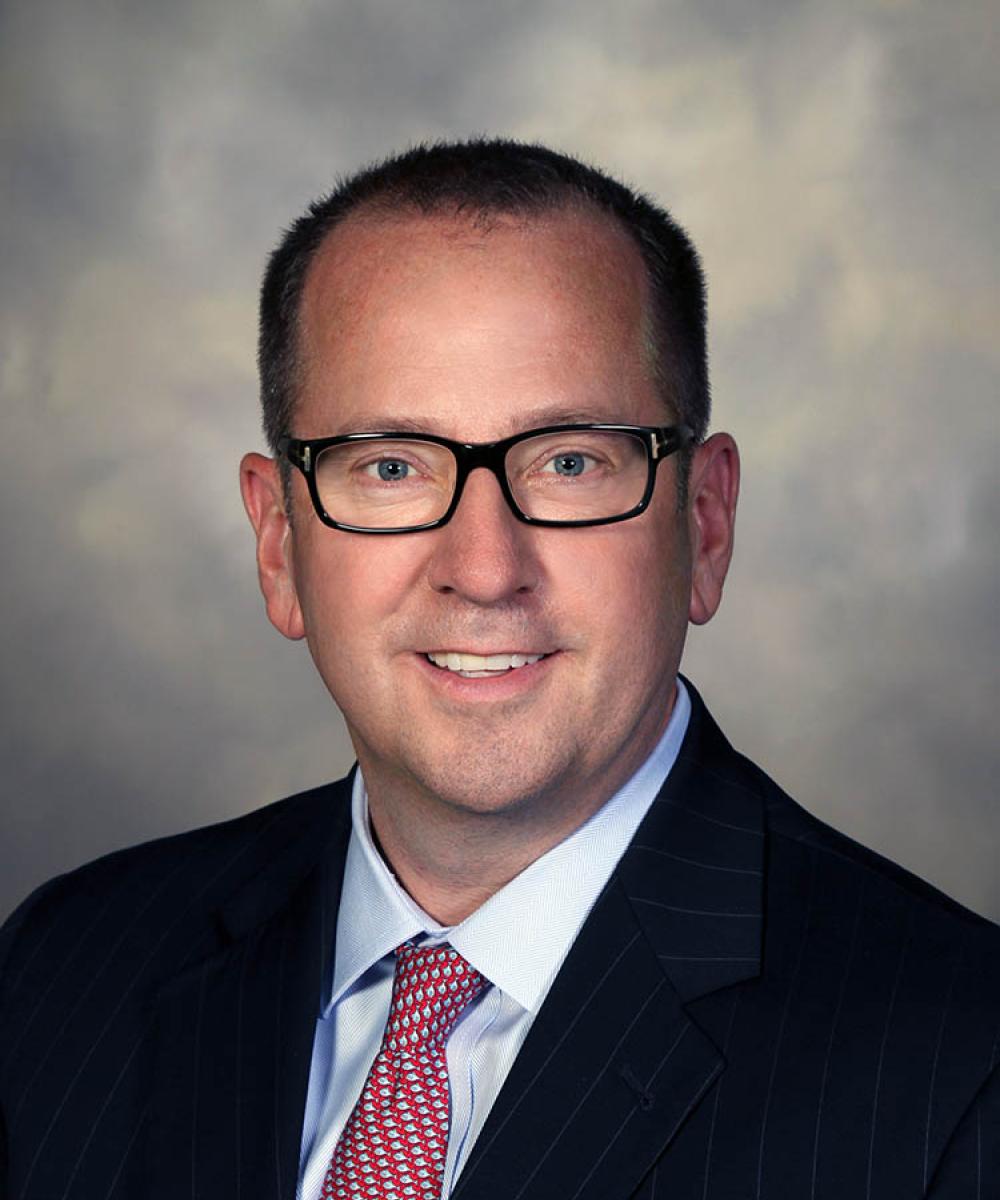 Headshot of Chris Swenson, wearing a white shirt, red tie, and dark blazer, with a mottled gray background