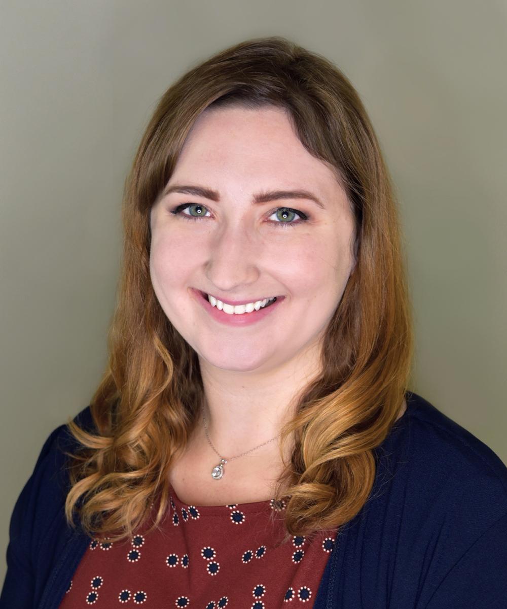Headshot of Claire Hamberg, wearing an orange shirt and blue cardigan, with a plain gray background