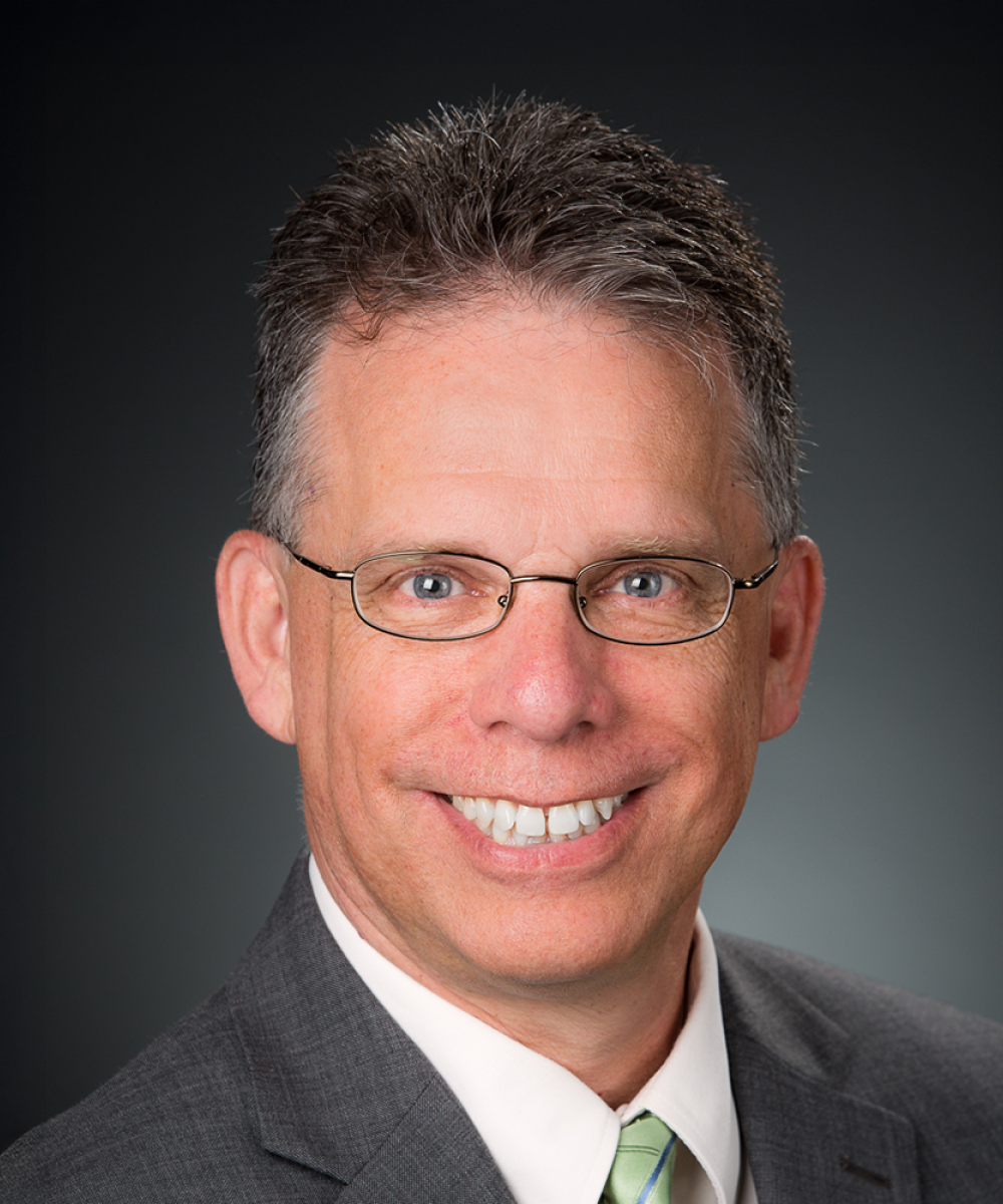 Headshot of Dave Kaplan, wearing a white shirt, green tie, and gray blazer, with a plain gray background