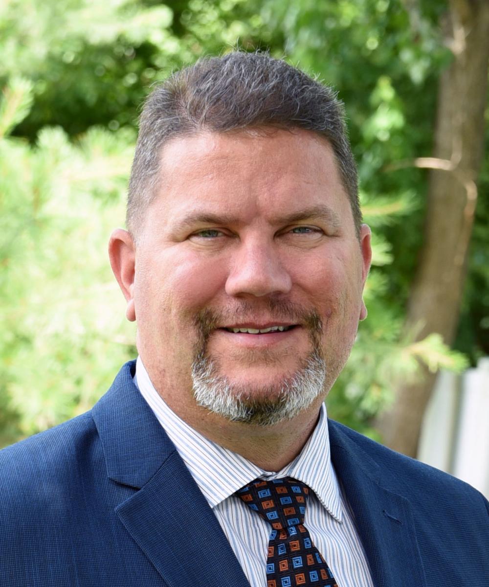 Headshot of Don Aulbert, wearing a striped shirt, patterned tie, and blue blazer, with a natural outdoor background
