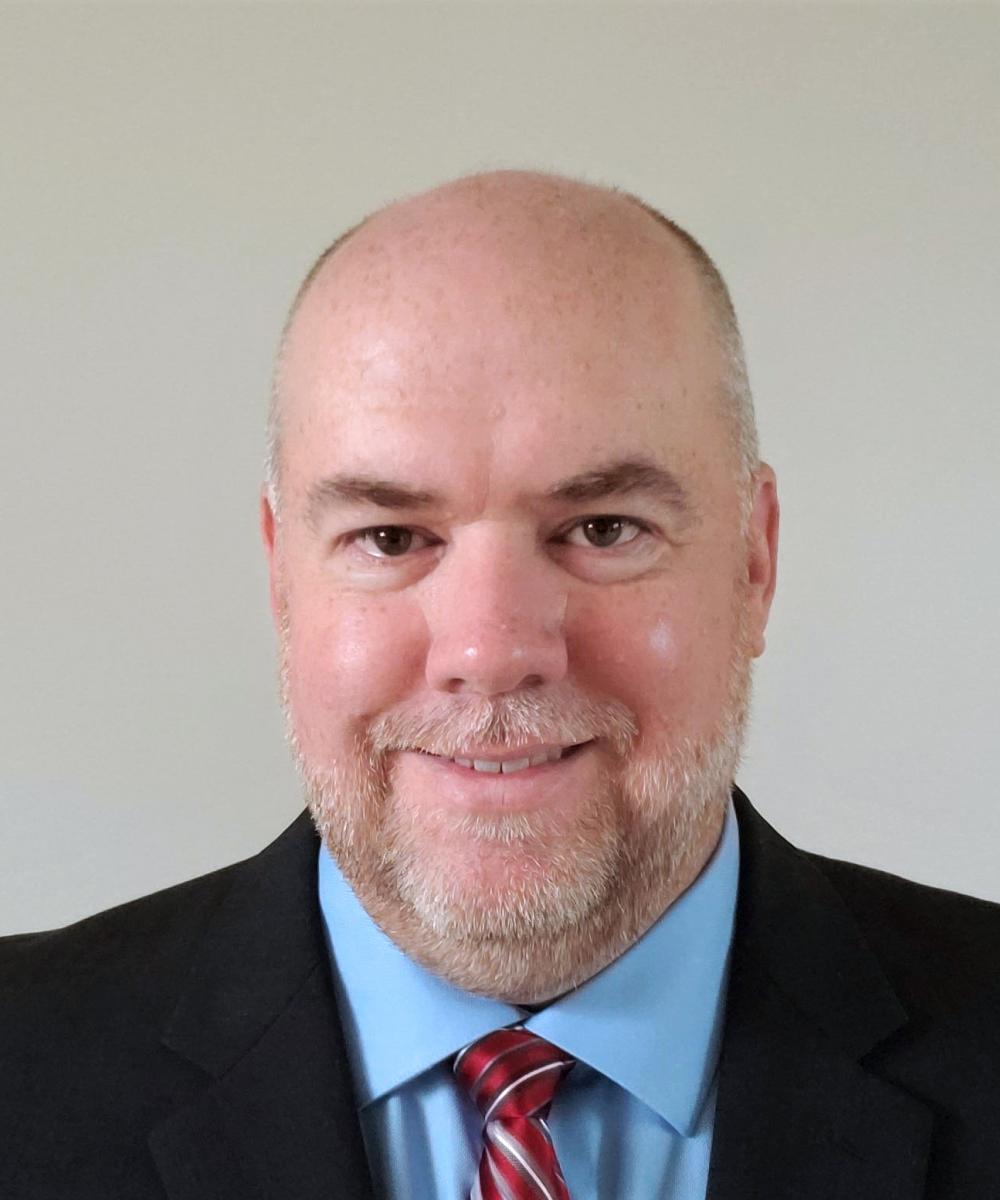 Headshot of John Howard, wearing a light blue shirt, red tie, and black blazer, with a plain gray background