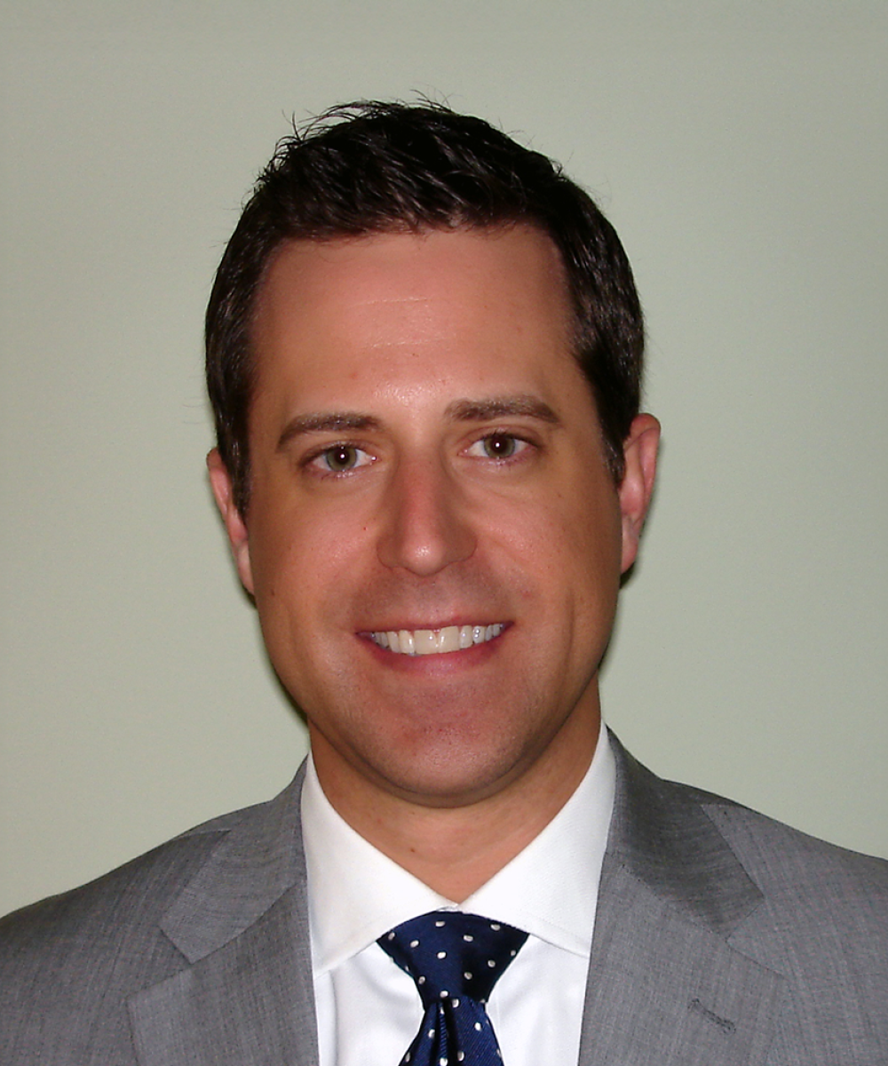Headshot of Mark Beckrich, wearing a white shirt, blue tie, and gray blazer, with a plain gray background