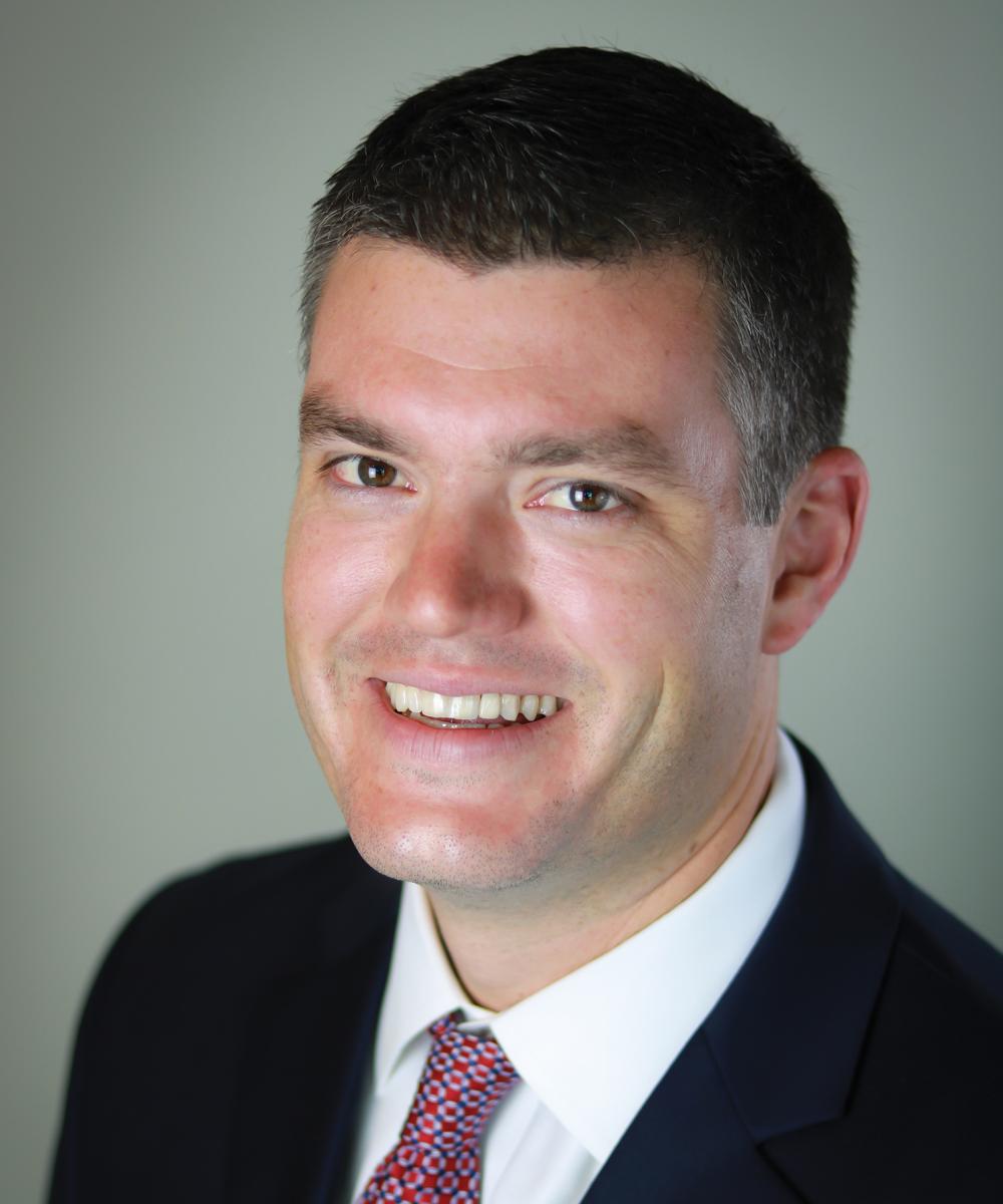 Headshot of Patrick Lodes, wearing a white shirt, red patterned tie, and dark blue blazer, with a plain gray background