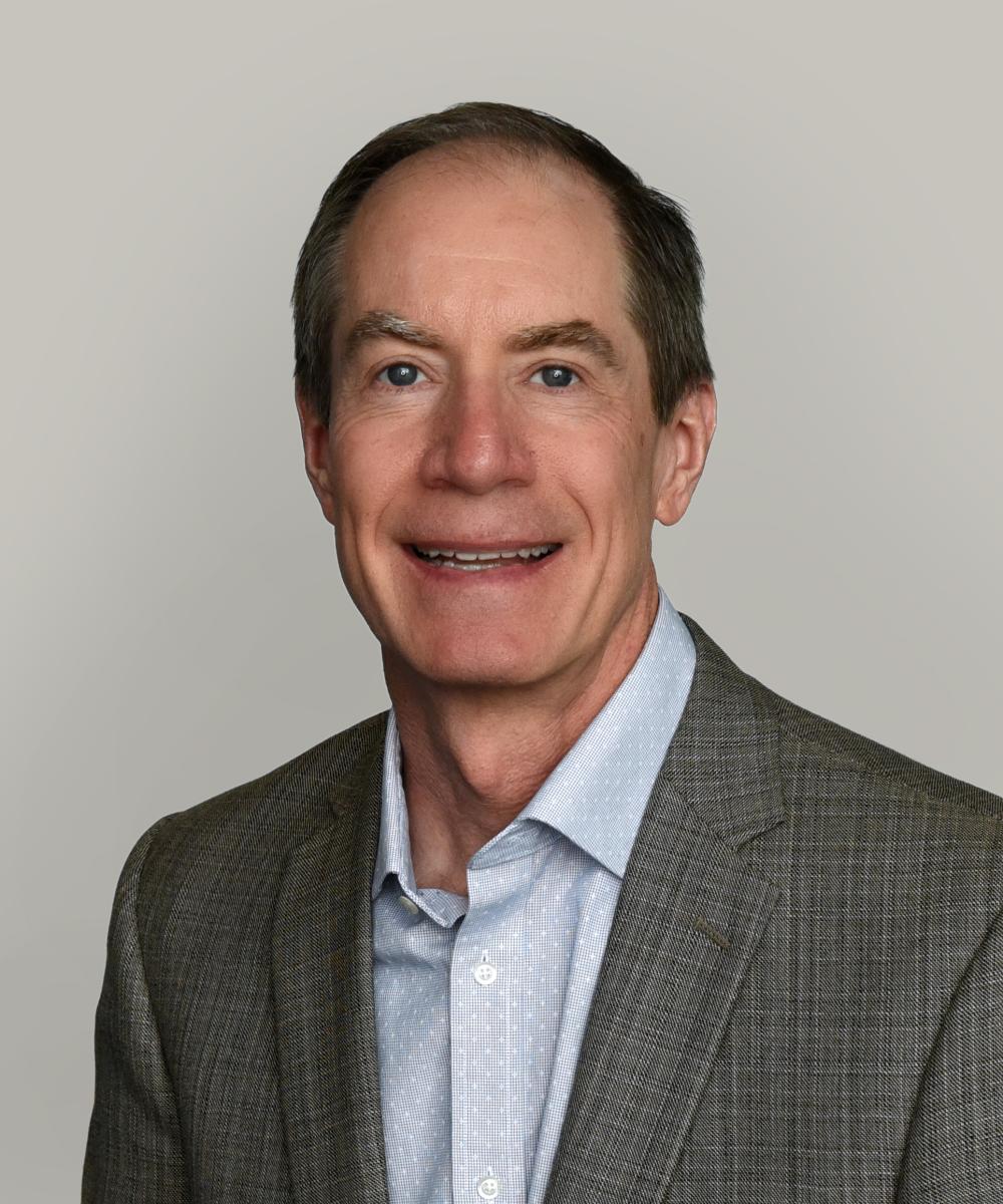 Headshot of Tom Gooden, wearing a light shirt and gray blazer, with a plain gray background
