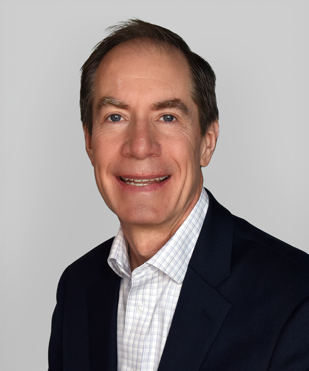 Headshot of Tom Gooden, wearing a white shirt and blue blazer, with a plain gray background